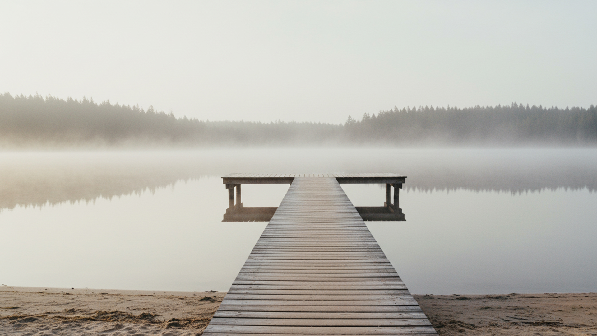 Serene wooden dock stretching into a misty, still lake surrounded by a forest at dawn.